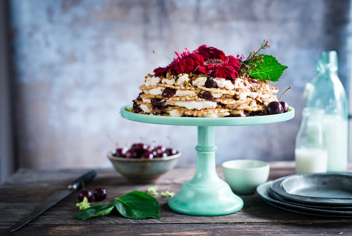 Tarta de capas finas con crema, cerezas y flores rojas, sobre un pie de tarta verde agua